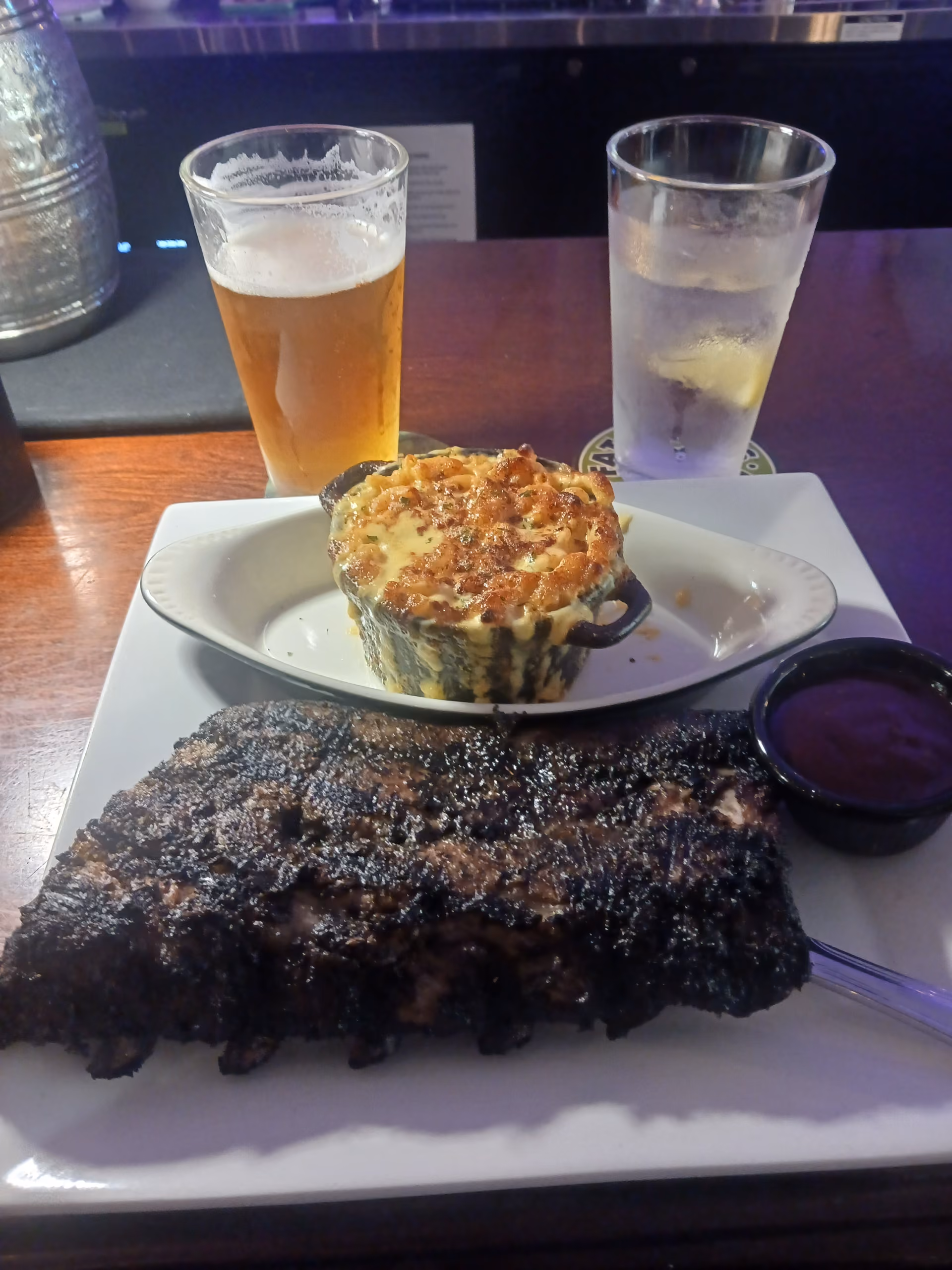 Plate with grilled ribs, macaroni and cheese, barbecue sauce, beer, and water with lemon on a wooden table.