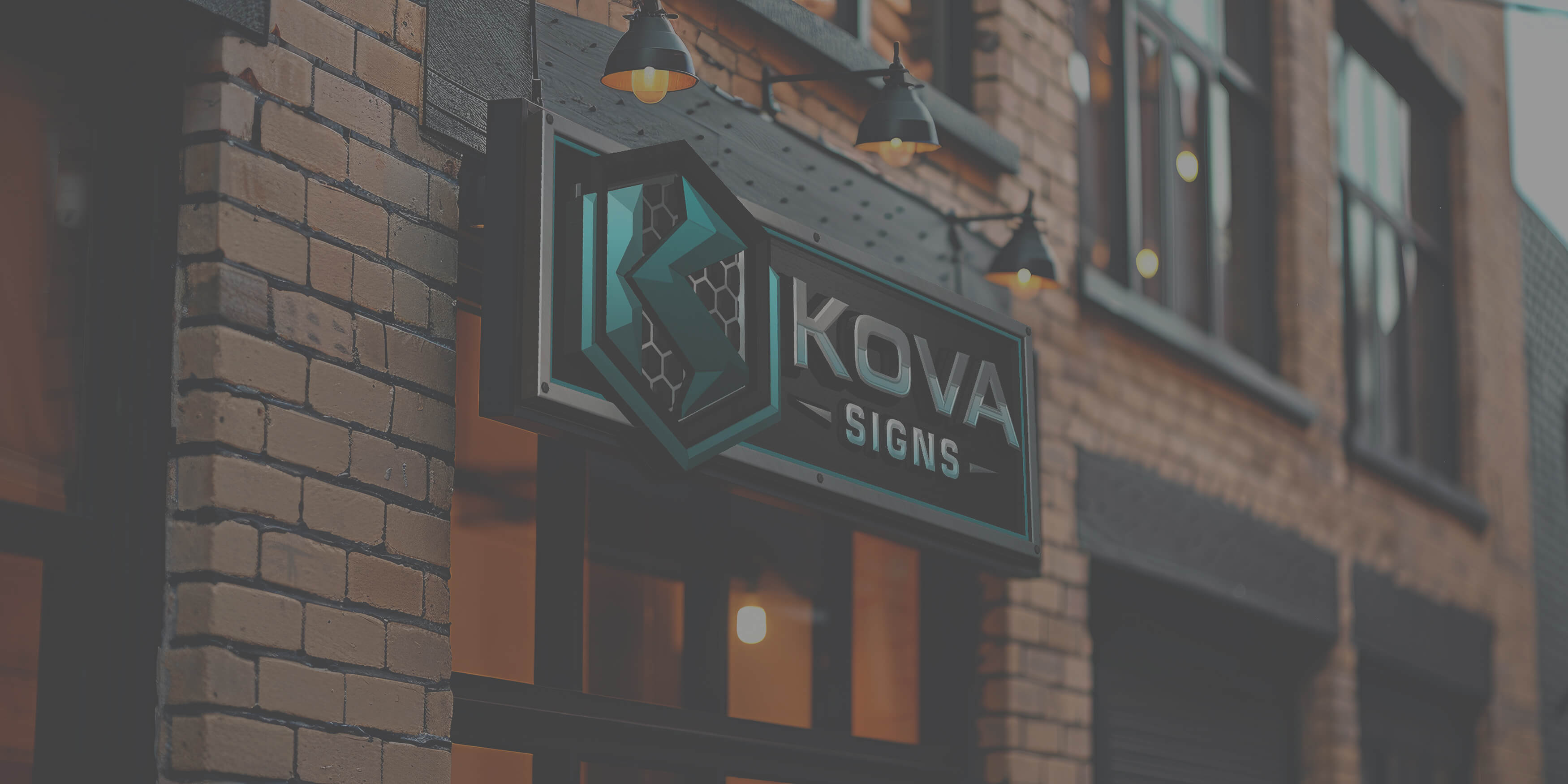 Black and teal KOVA SIGNS store sign mounted on a brick building under hanging lamps.