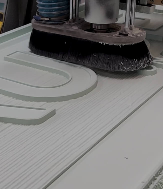 Close-up of a CNC machine carving raised letters into a textured white wood surface.