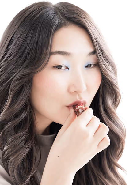 Close-up of a woman with long wavy dark hair and light blue eyeshadow eating a small piece of food held near her lips with her right hand.