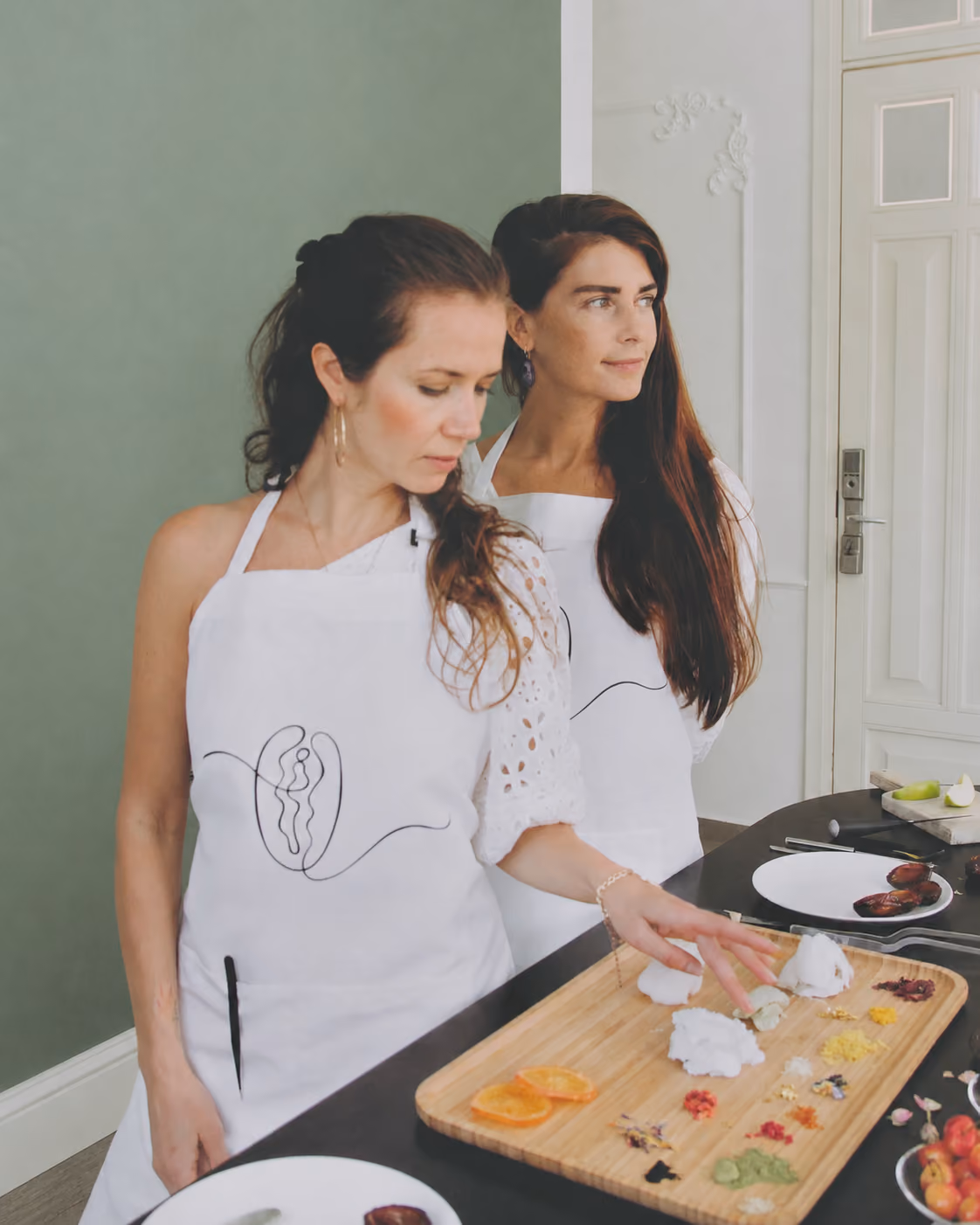 Two women wearing white aprons standing in a kitchen. One woman is reaching towards a wooden cutting board with various colorful small ingredients and dollops of white substance arranged neatly on it. The other woman looks to the side. The countertop has plates with more ingredients and kitchen utensils.