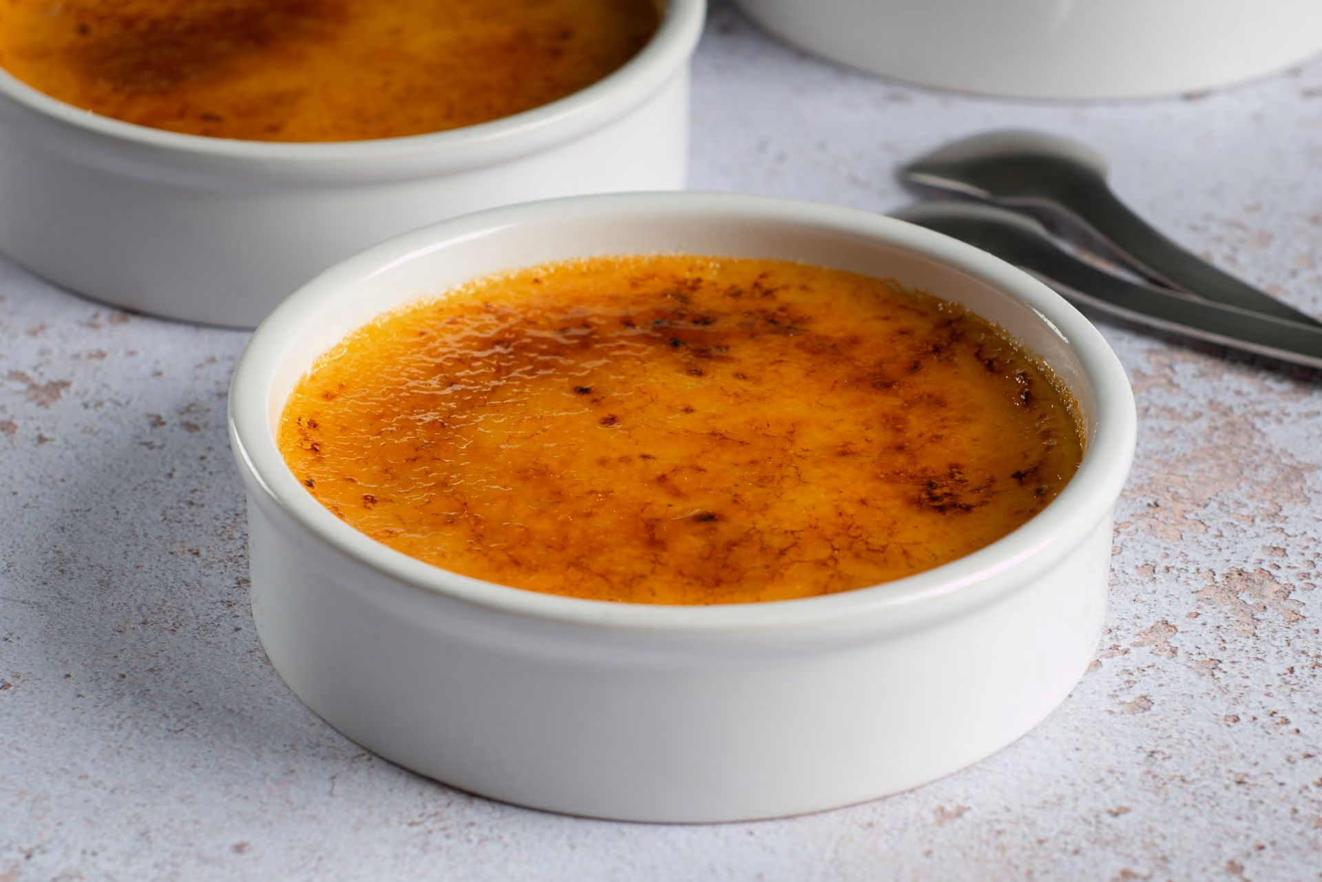 Close-up of crème brûlée with caramelized sugar crust in white ramekin on a textured surface.