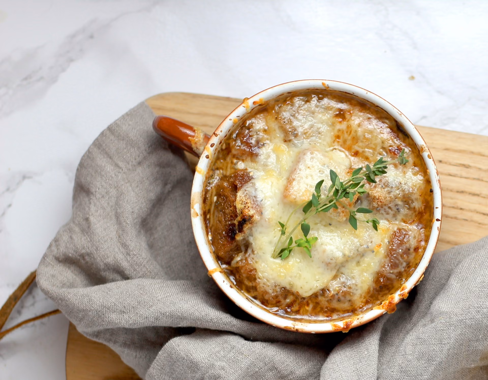 Baked French onion soup with melted cheese and herbs in a brown handled bowl on cloth and wood.