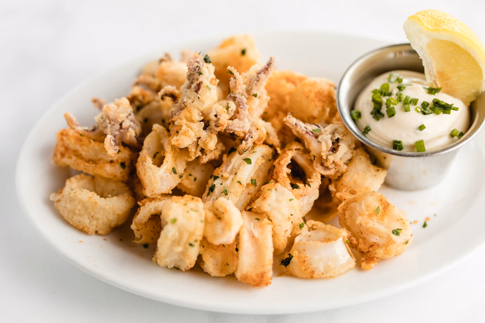 Plate of golden fried calamari with a lemon wedge and creamy dipping sauce topped with chopped herbs.