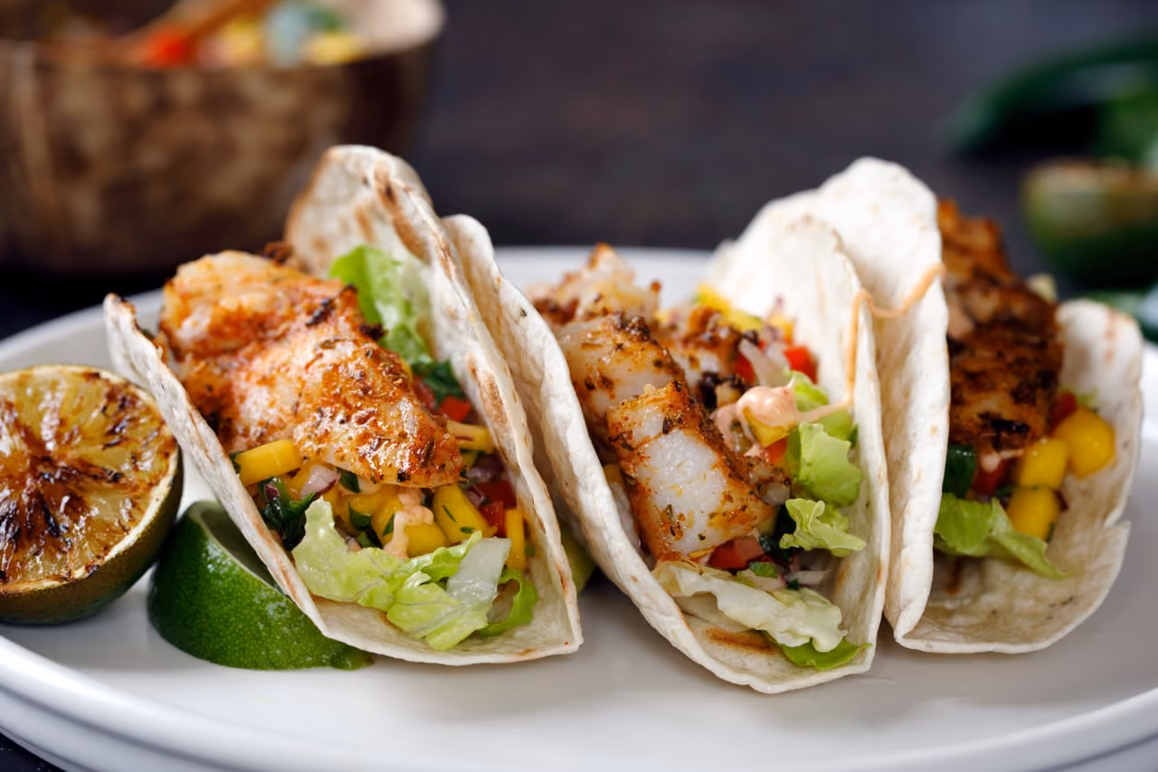 Three grilled fish tacos with lettuce, mango salsa, and sauce on a white plate with grilled lime halves.