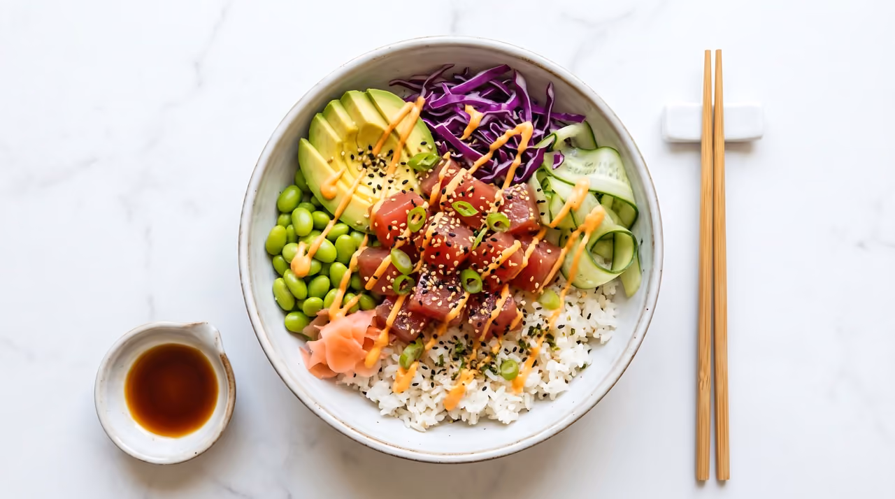 Bowl of poke with tuna, rice, avocado, edamame, cucumber, purple cabbage, and sauce, with chopsticks and soy sauce.