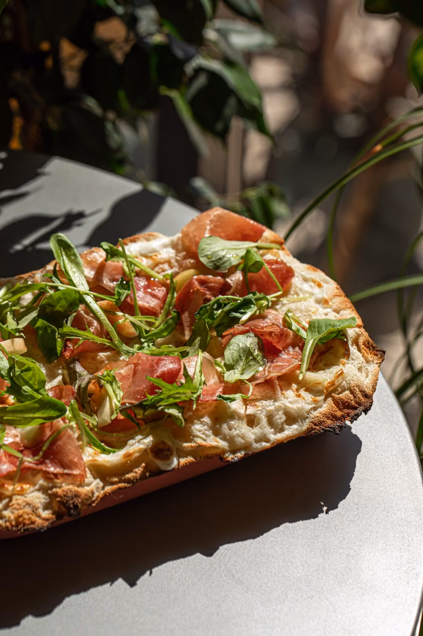 Flatbread pizza topped with greens and slices of prosciutto on a round table in natural light.