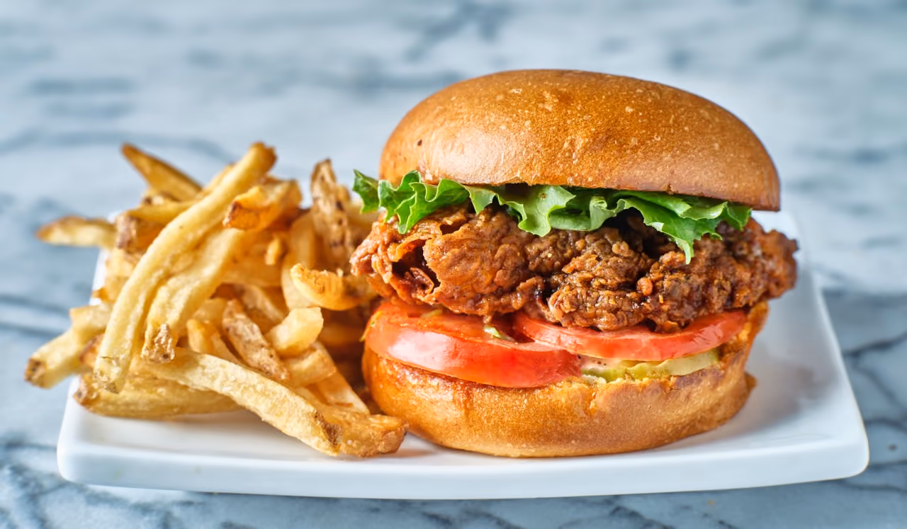 Crispy fried chicken sandwich with lettuce and tomato, served with French fries on a white plate.