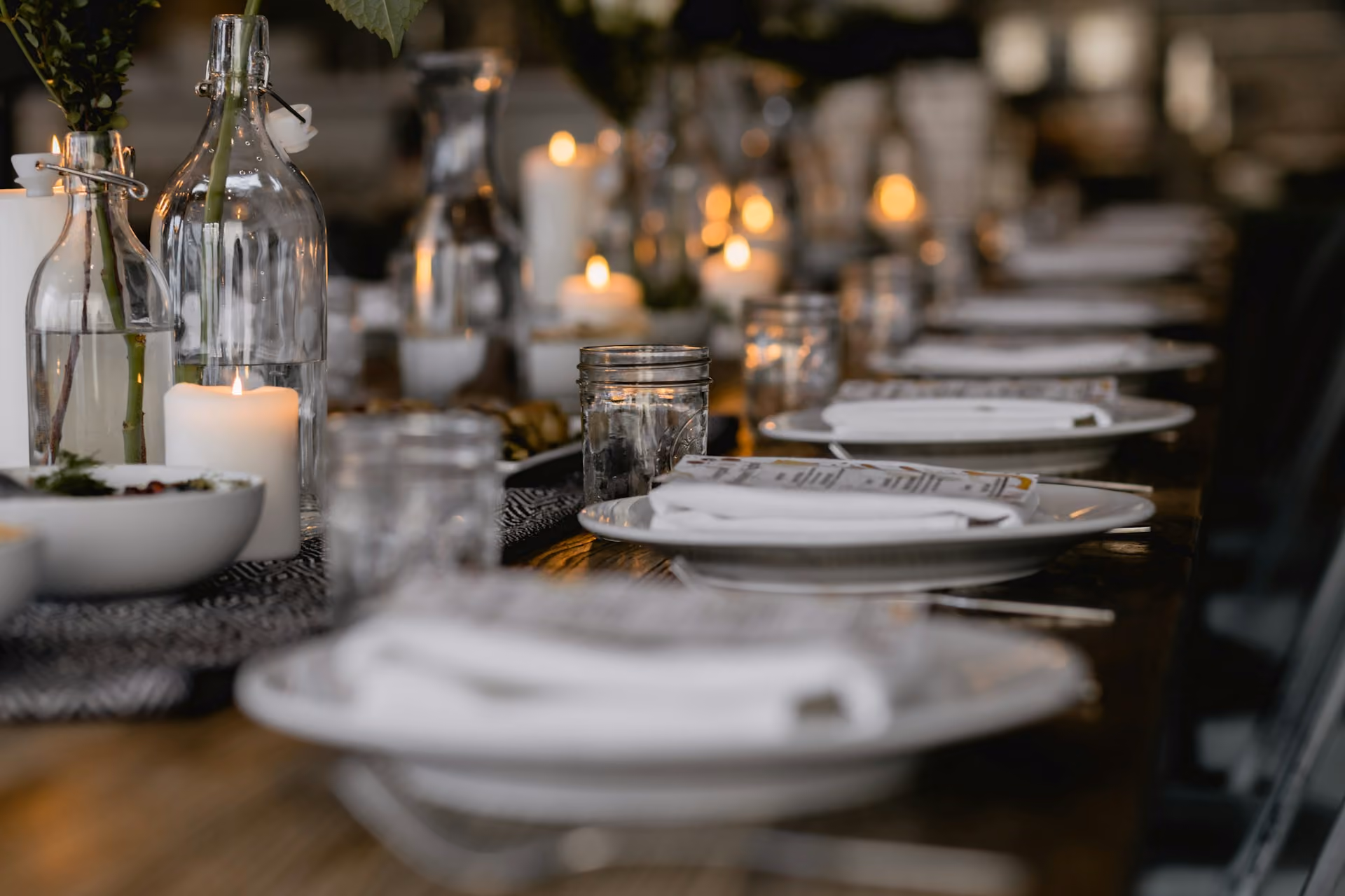 Long wooden table set with plates, napkins, glass jars, candles, and glass vases with flowers.