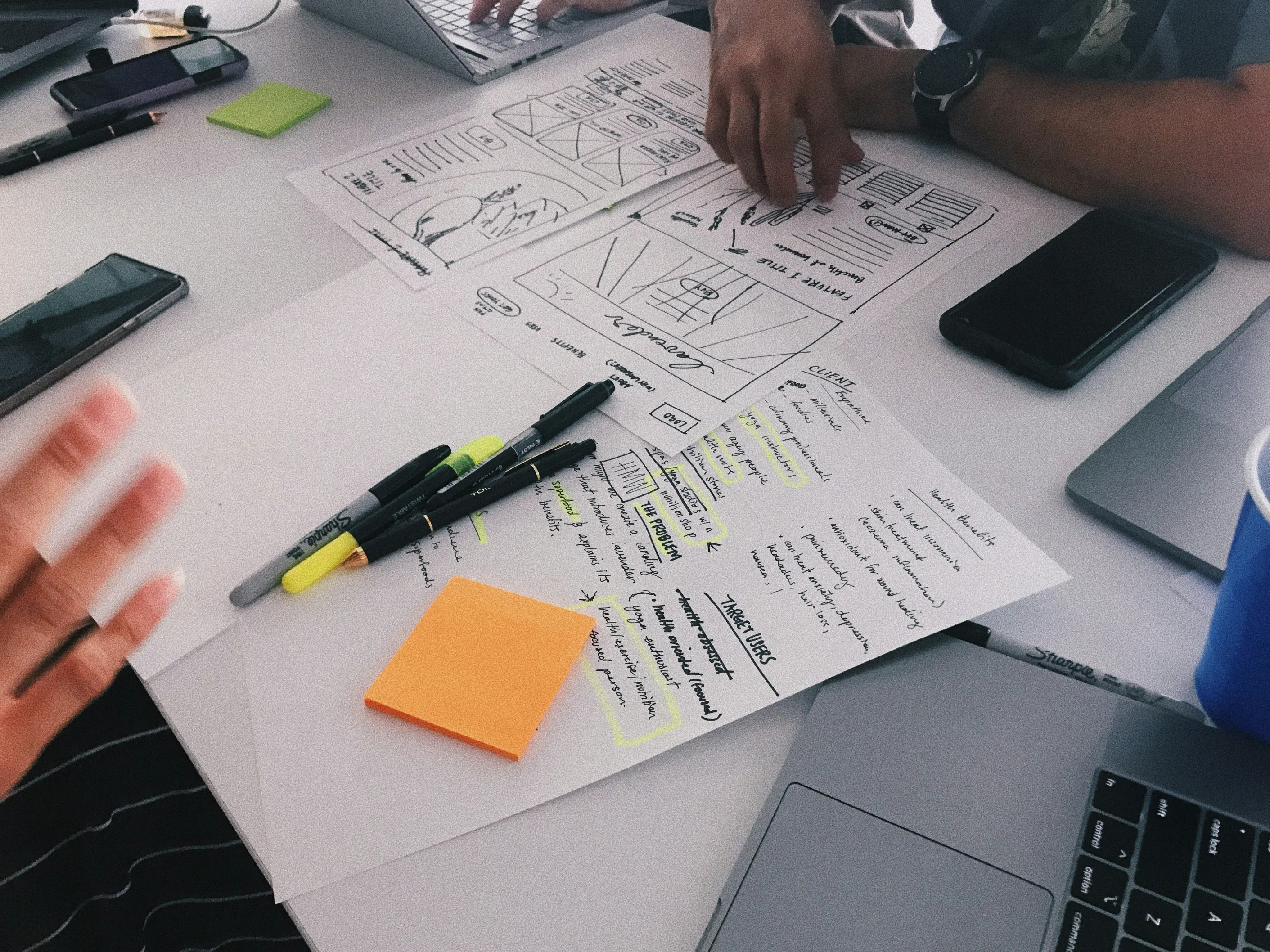 A designer sketching app wireframes on paper beside sticky notes and a laptop
