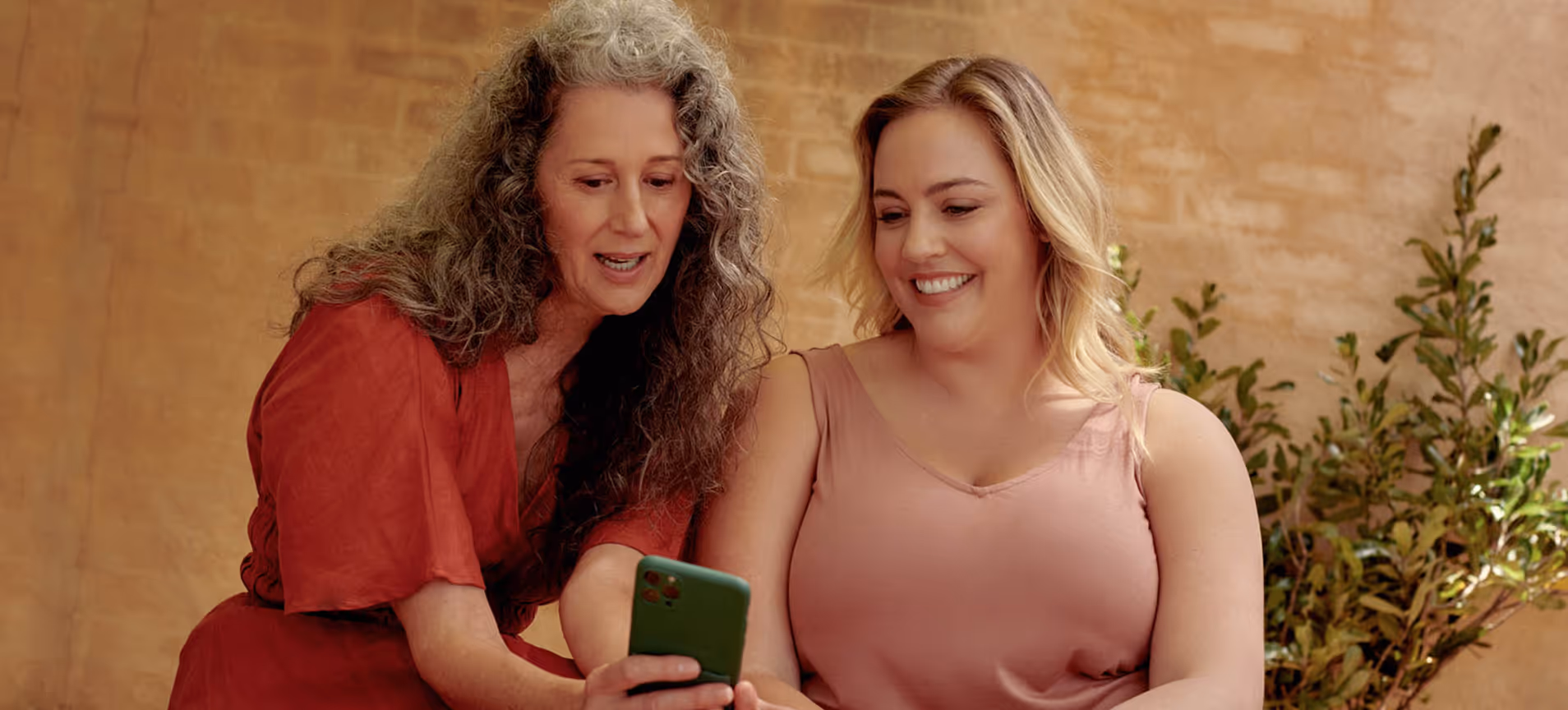 Two women smiling together over a phone, supporting each other
