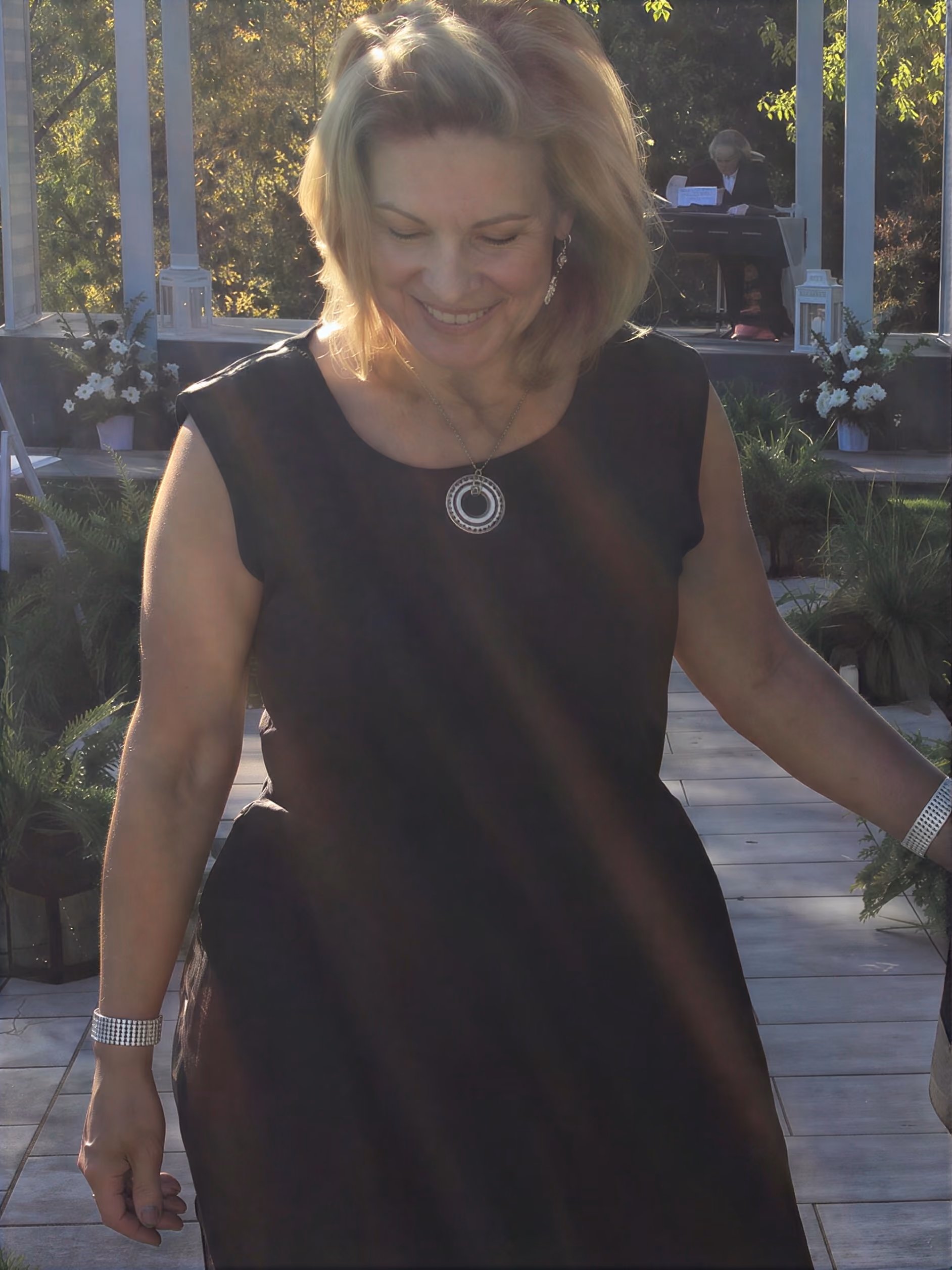Smiling woman in black dress outdoors with sun rays and plants around her.