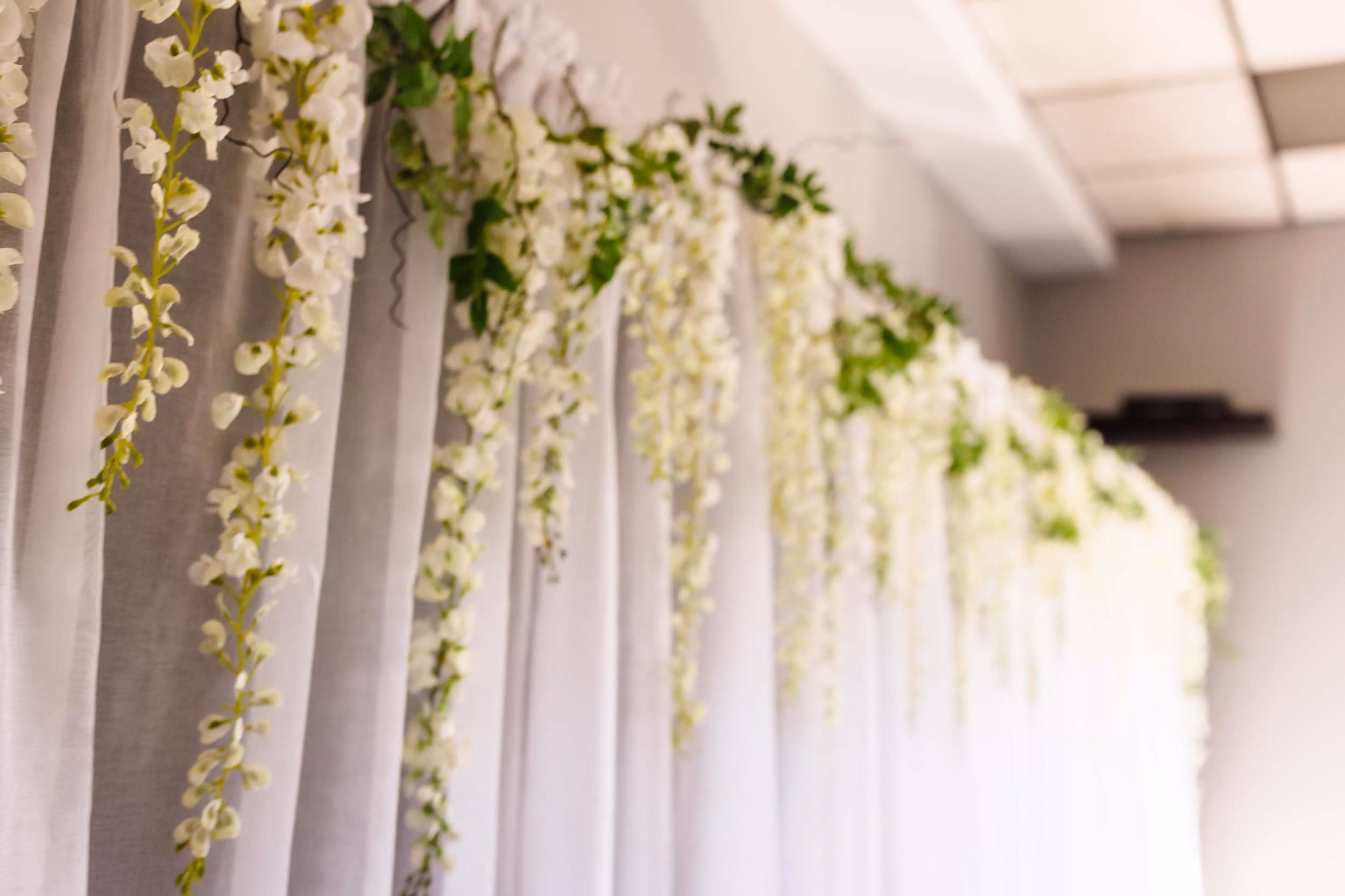 White hanging flowers with green leaves draped along white sheer curtains in a room.