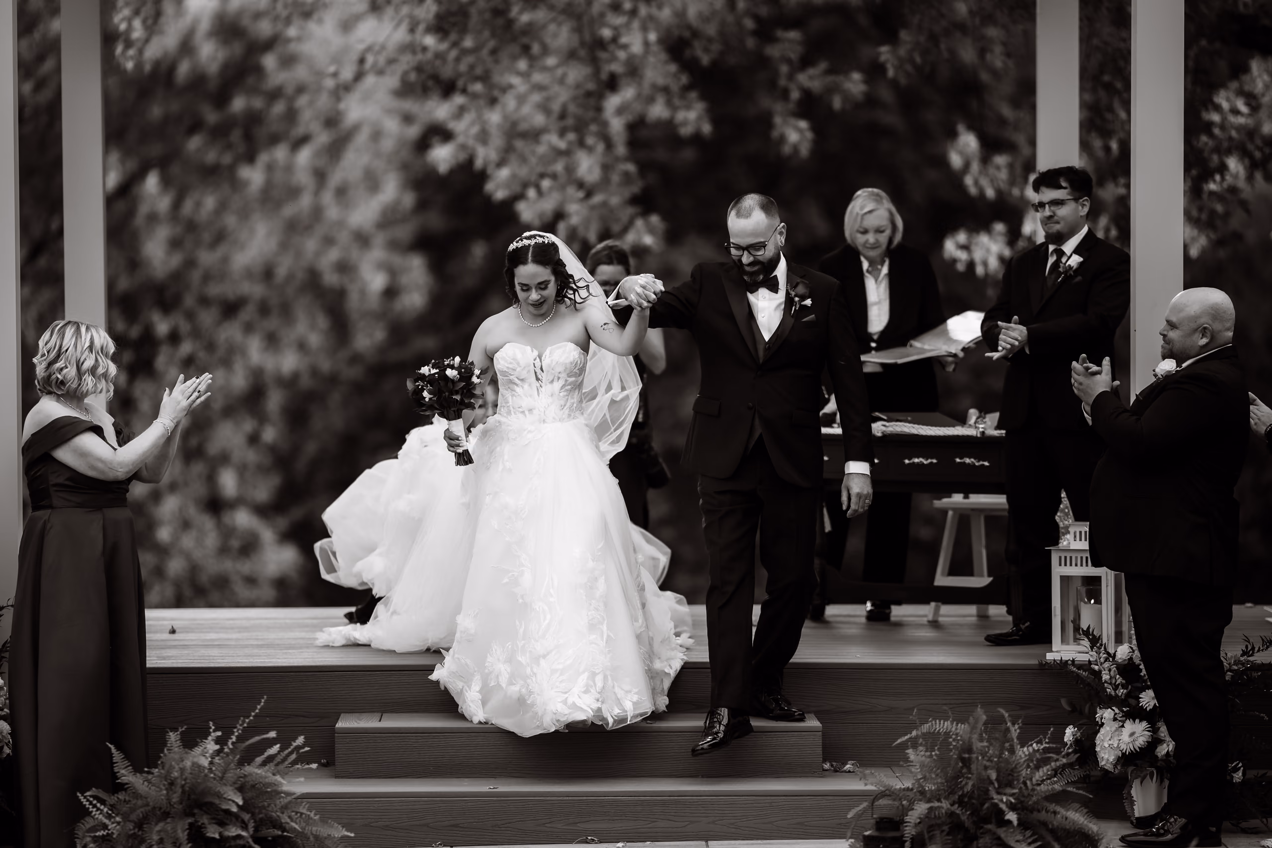 Bride and groom hold hands while walking down steps amid applauding guests at outdoor wedding ceremony.