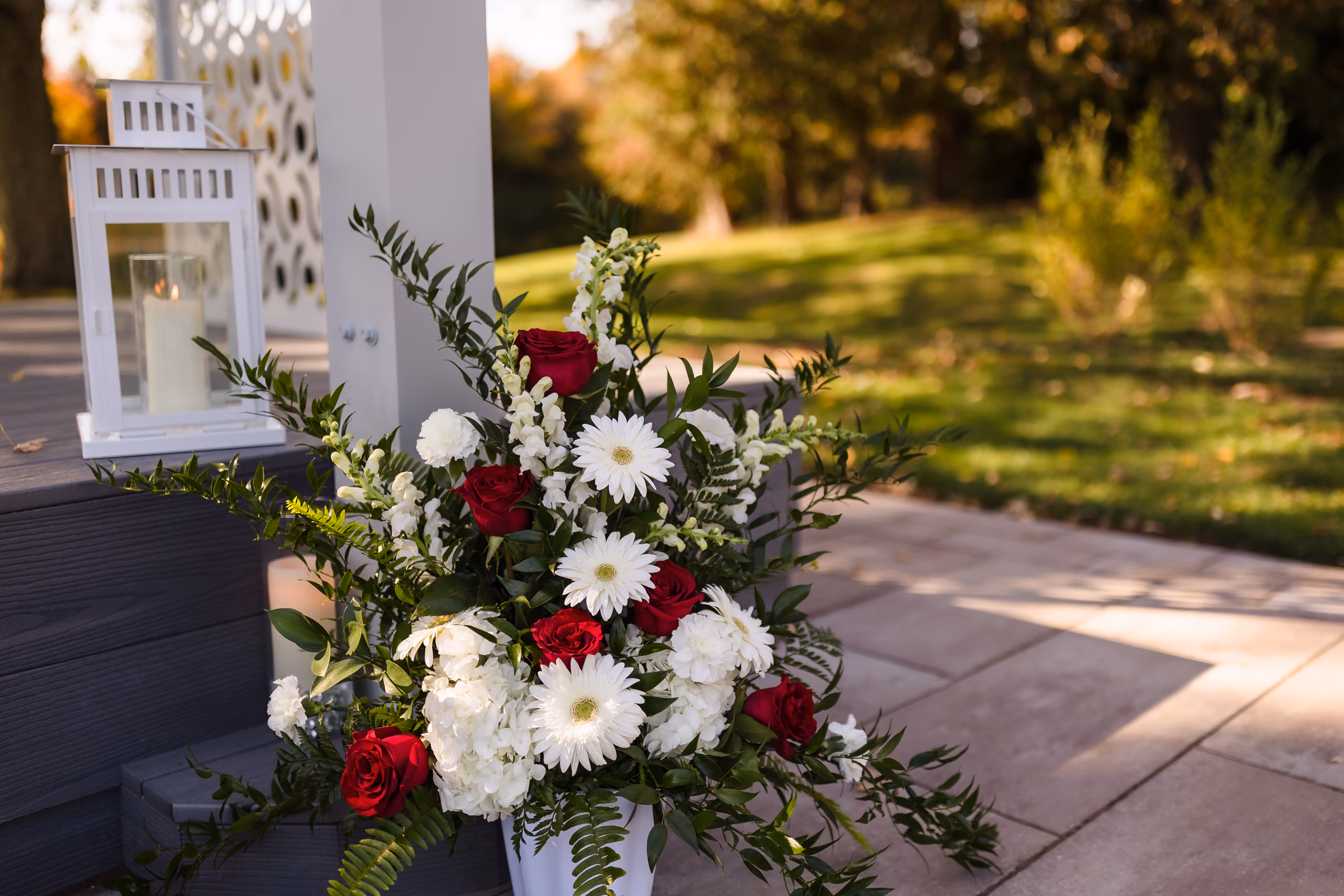 Bouquet of red roses and white flowers with greenery next to a white candle lantern outdoors.