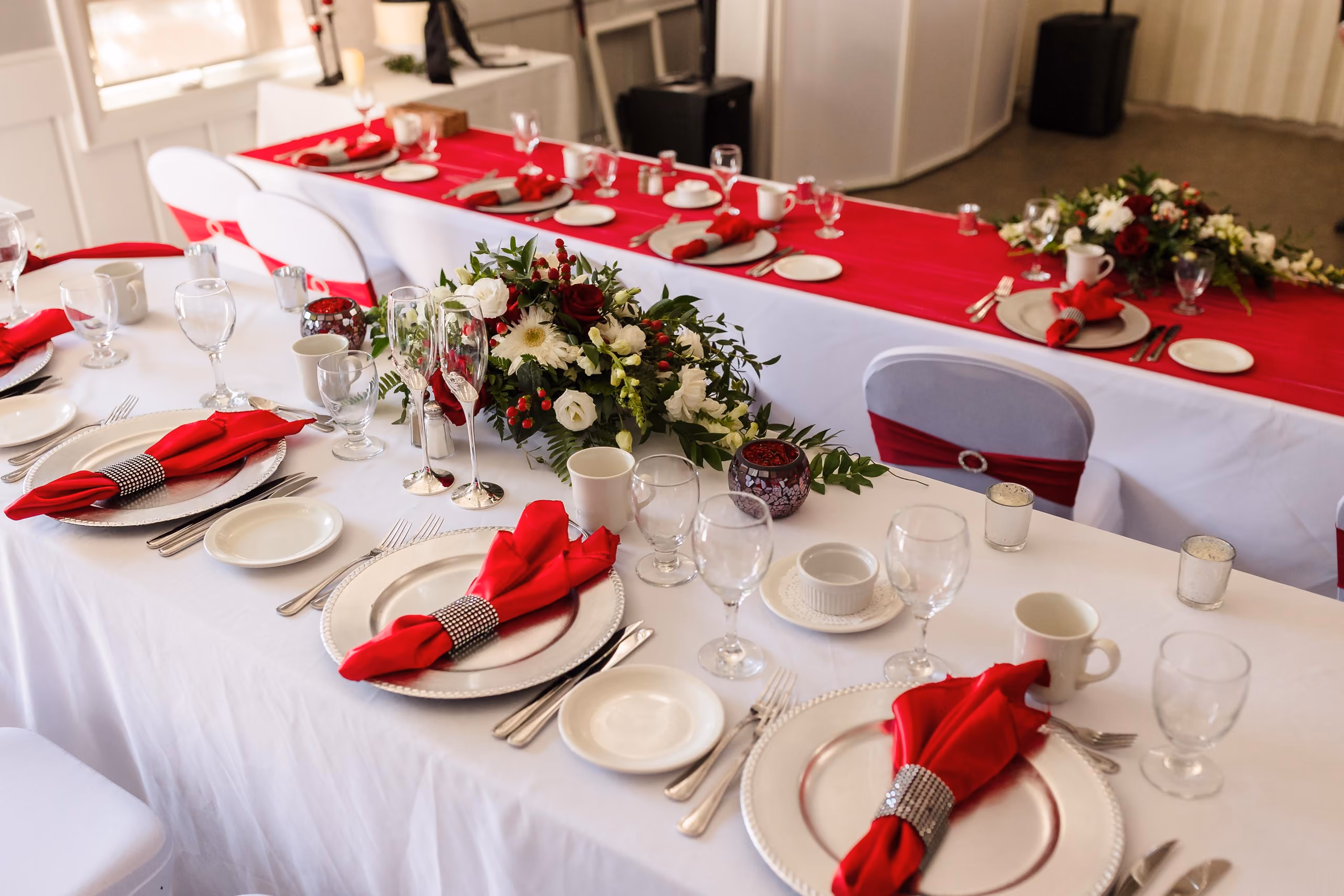 Table set with white cloths, red napkins, glassware, and floral centerpieces for a formal event.