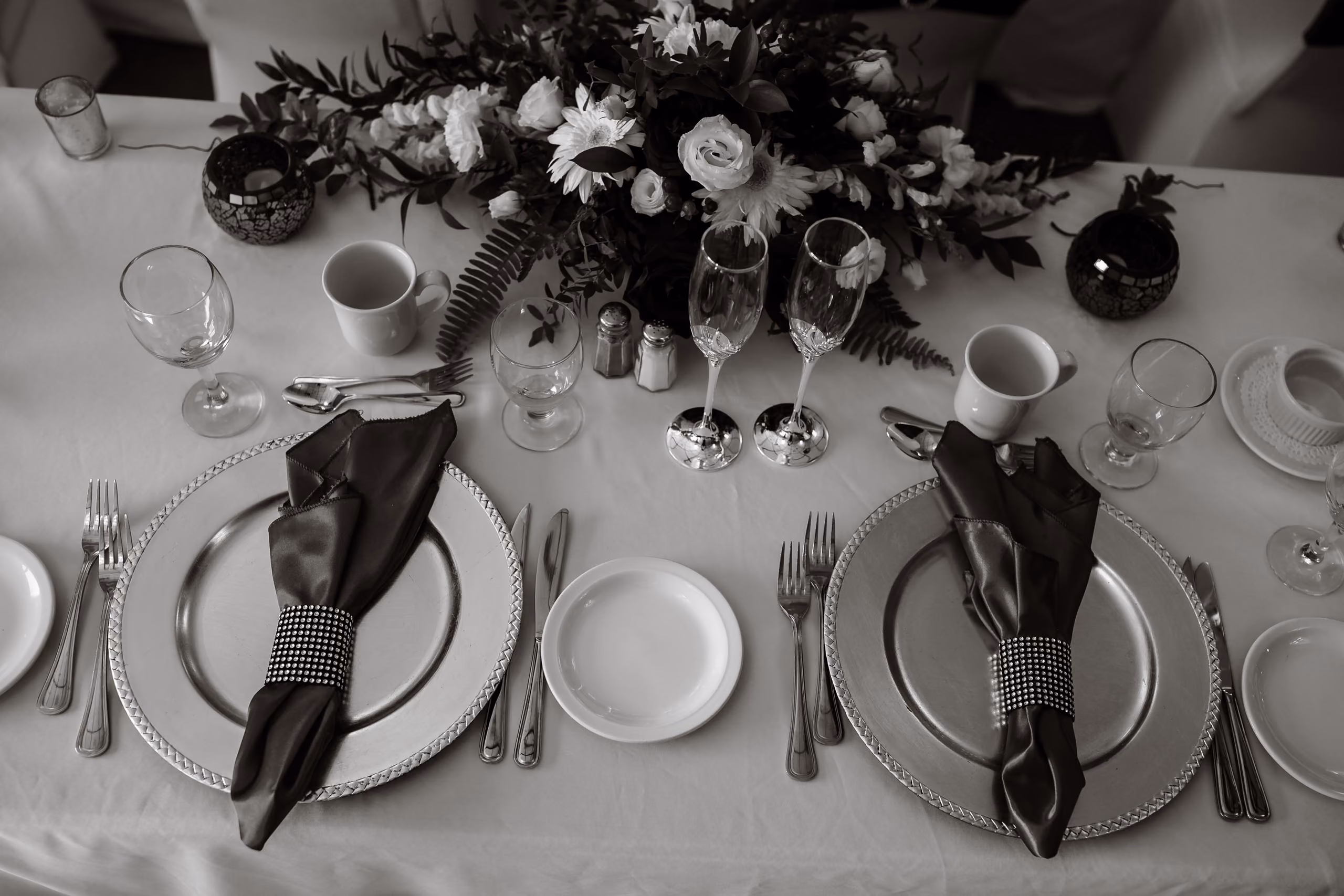 Elegant table set for two with floral centerpiece, silver plates, black napkins, glasses, and cutlery.