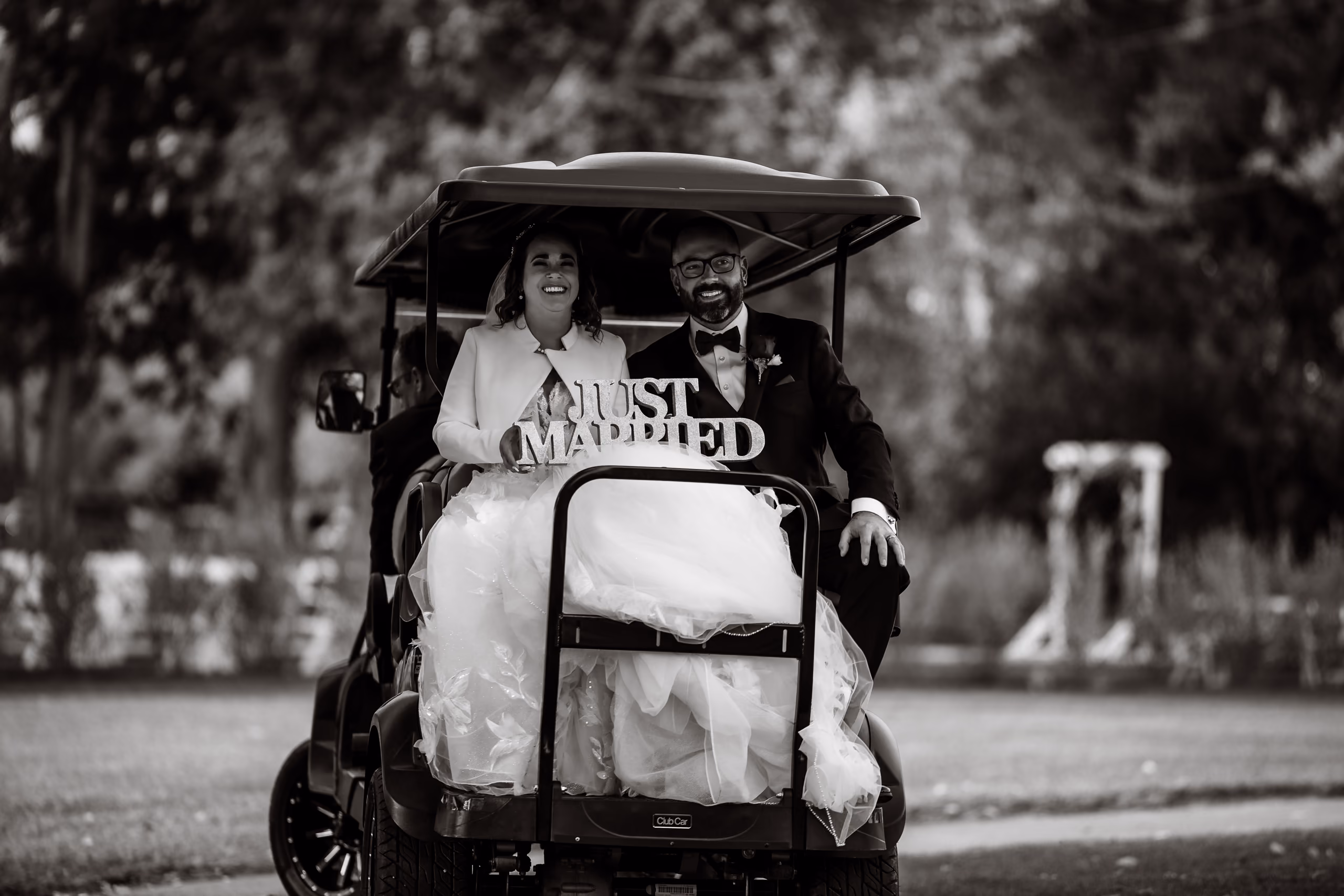 Bride and groom smiling on a golf cart holding a 'Just Married' sign in black and white photo.