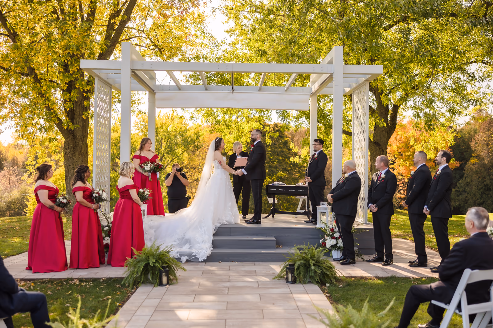 Bride and groom hold hands under white pergola with bridal party in red dresses and black suits.