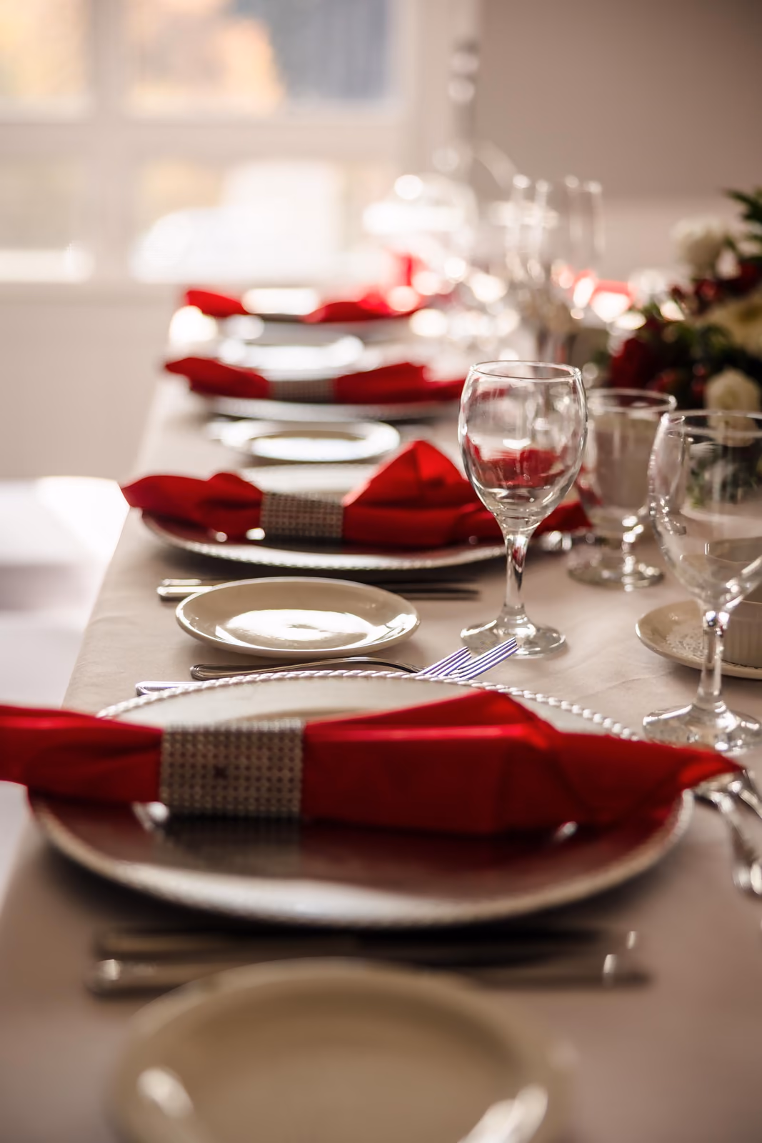 Elegant dining table set with silver plates, red napkins, wine glasses, and cutlery on a white tablecloth.