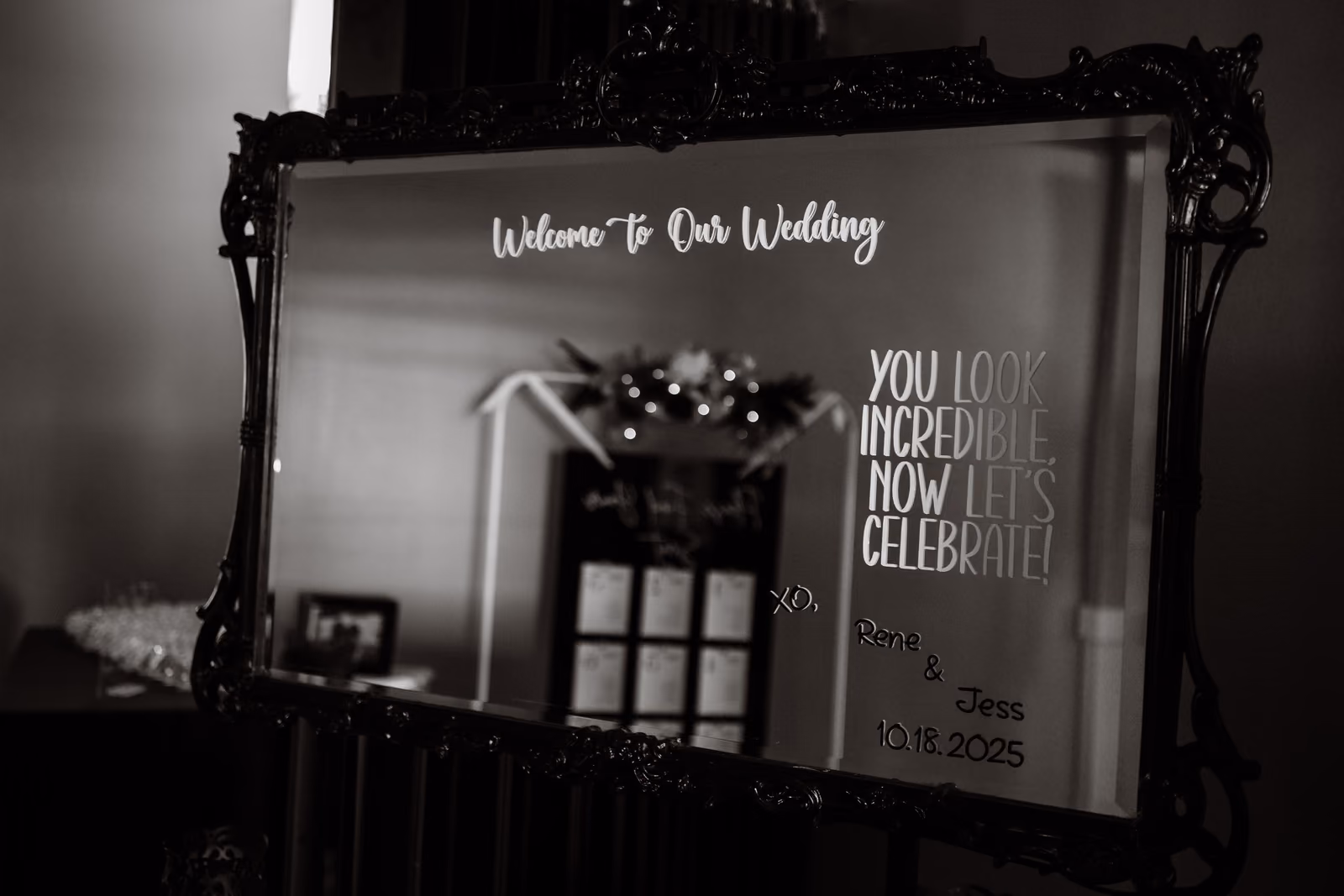 Ornate mirror with wedding welcome message and date 10.18.2025 in a dimly lit room.