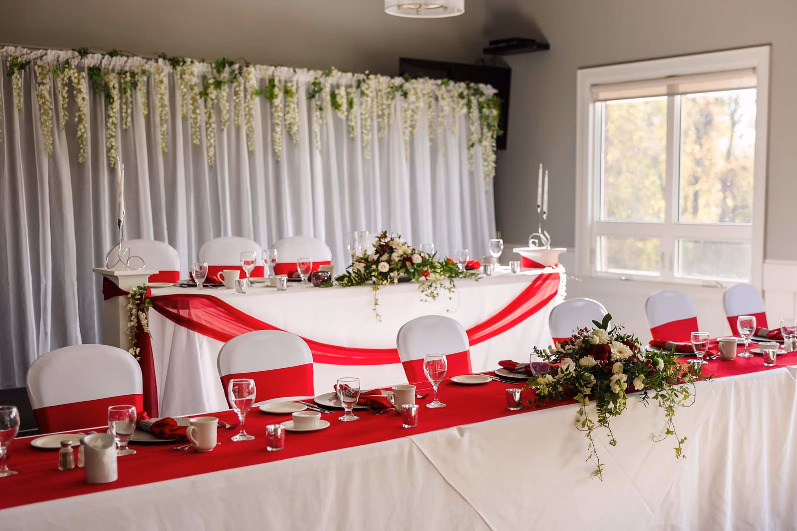 Wedding head table with white and red decorations, floral centerpieces, and glassware in a bright room.