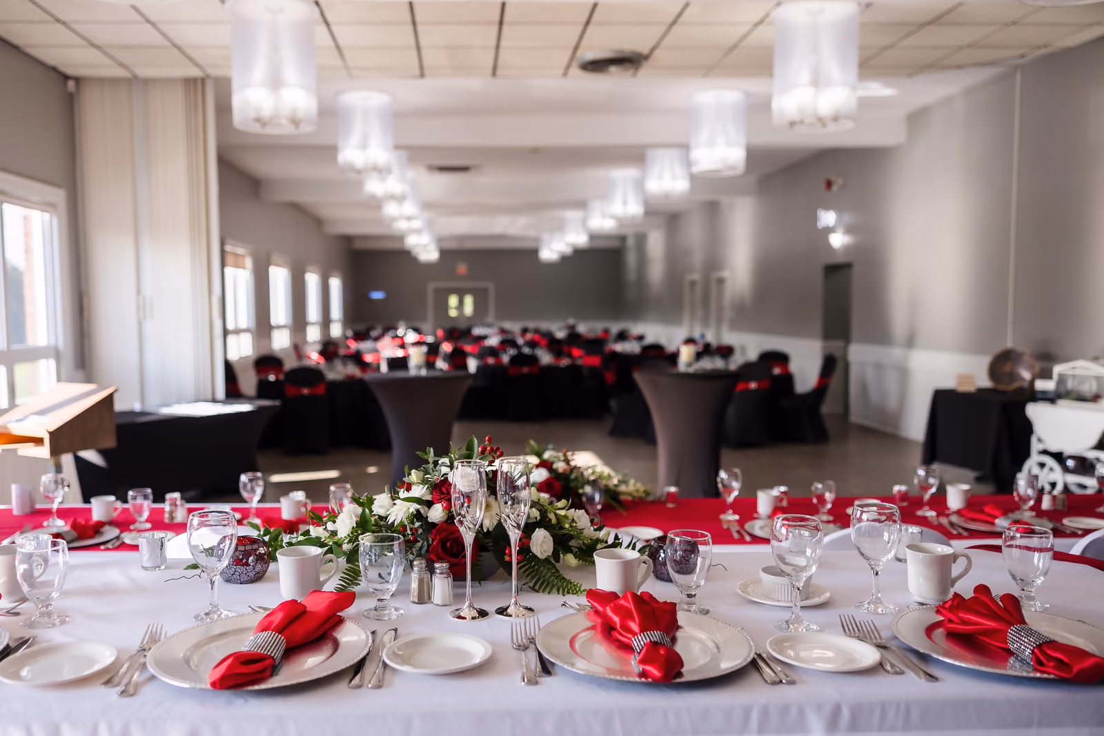 Elegant banquet hall with tables set in white, red napkins, silver plates, and floral centerpieces.