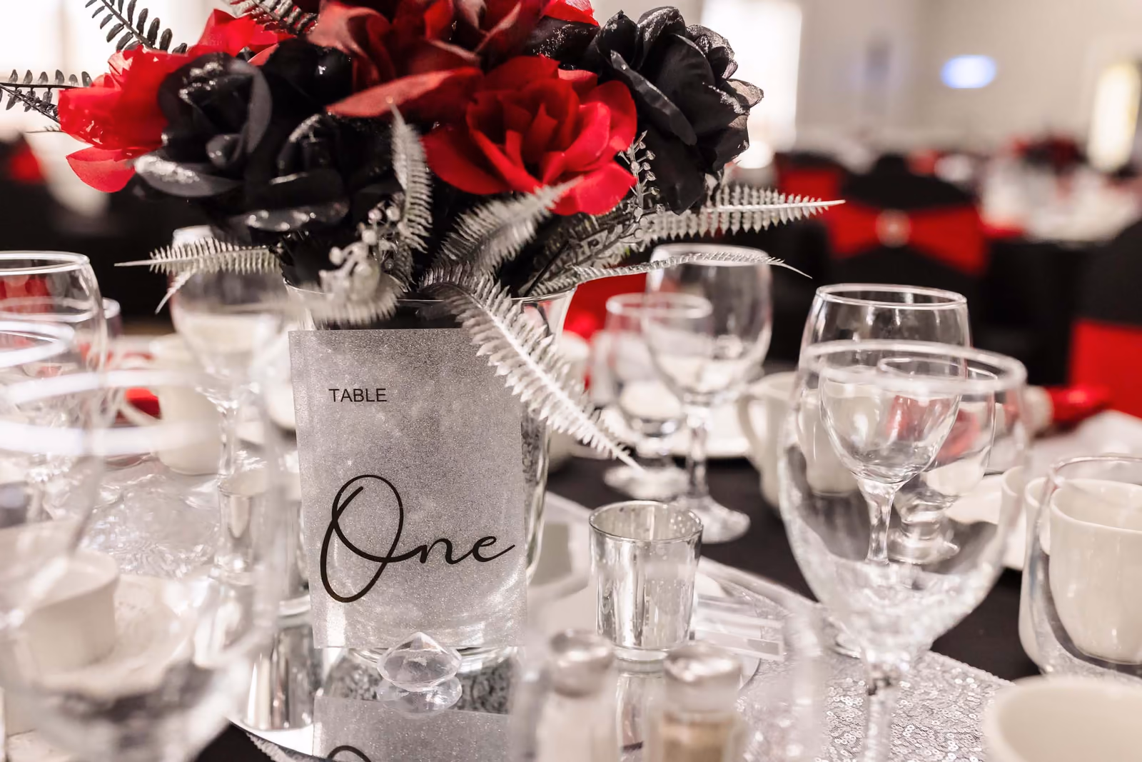 Table one centerpiece with red and black roses, glassware, and white cups on a decorated table.