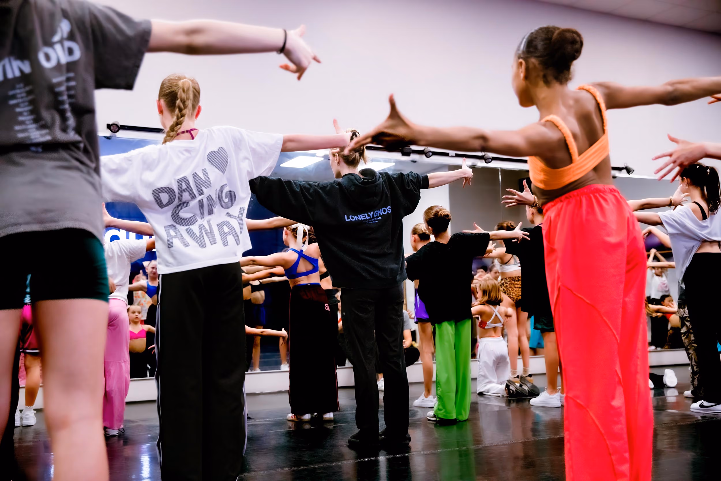 A group of diverse teens dancing in a studio with arms outstretched, practicing in front of mirrors.