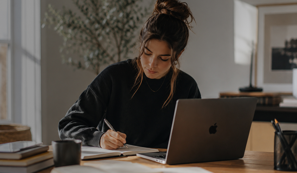 Young woman with hair in a bun writing in a notebook at a desk with an open laptop and books.