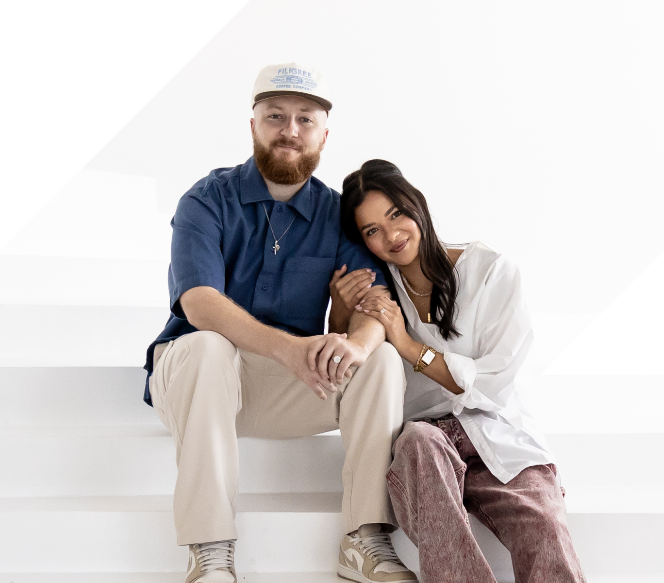 Couple sitting on white stairs, woman leaning on man’s shoulder smiling at camera.