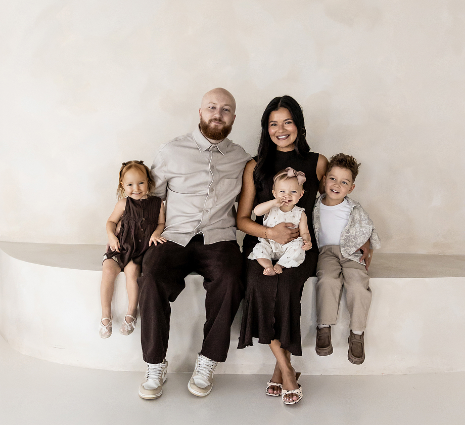 Smiling family of five seated on a curved white bench against a beige wall.