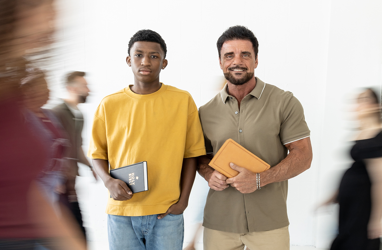 Two men standing against a white wall holding books, with blurred people walking past them.