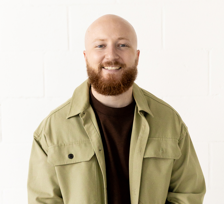 Smiling bald man with red beard wearing an olive green jacket and dark shirt against a white background.
