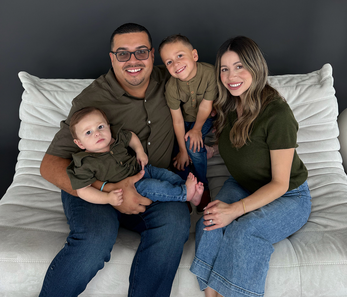 Smiling family of four wearing green shirts and jeans sitting on a white sofa against a dark background.