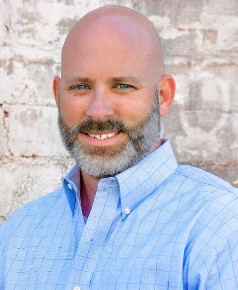 Headshot of David Pratt, Executive Vice President & General Counsel at SummitView Holdings, wearing a light blue checked shirt in front of a whitewashed brick wall.