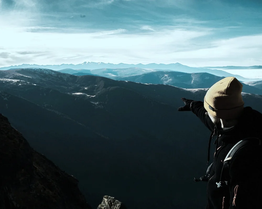 A hiker in a beanie and backpack stands on a mountain summit pointing toward a distant peak, representing SummitView Holdings' team development and leadership.