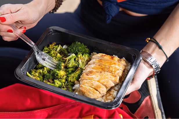 Person holding a container with cooked broccoli and sliced grilled chicken over rice.