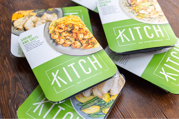 Stack of green and white Kitch meal packages featuring shrimp and grits on a wooden table.