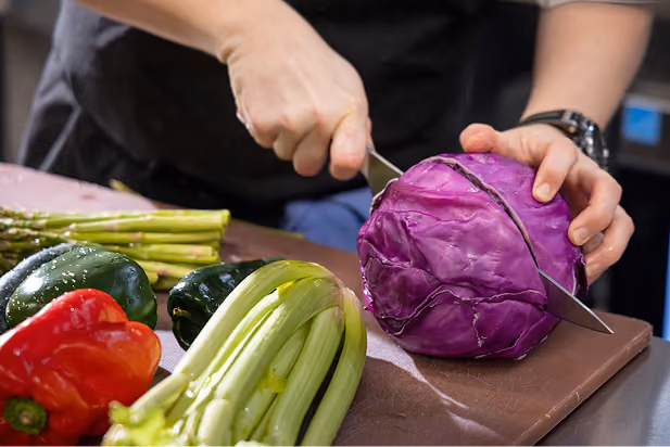 Person cutting a purple cabbage on a cutting board with fresh vegetables nearby.