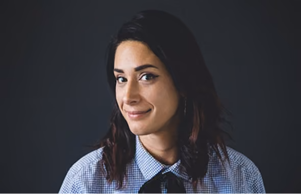 Woman with dark hair wearing a checkered shirt and smiling softly against a dark background.
