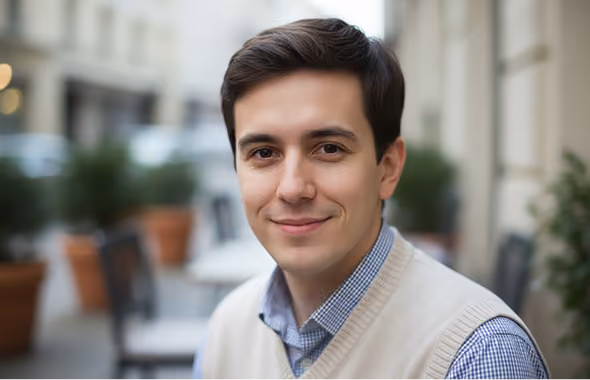 Young man with dark hair smiling softly, wearing a beige sweater vest and checked shirt outdoors.