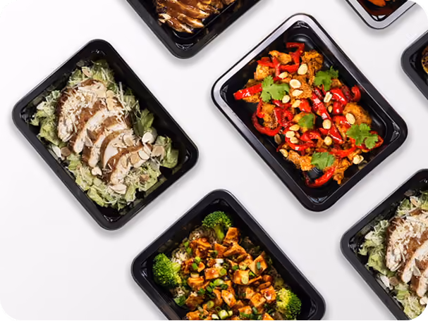 Black trays with a variety of prepared meals including chicken salad and vegetable dishes on a white surface.