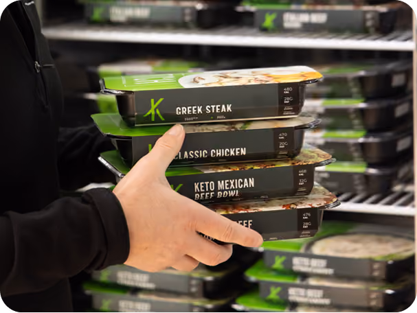 Hand holding stacked packaged Kitch meals labeled Greek Steak, Classic Chicken, Keto Mexican Beef Bowl.