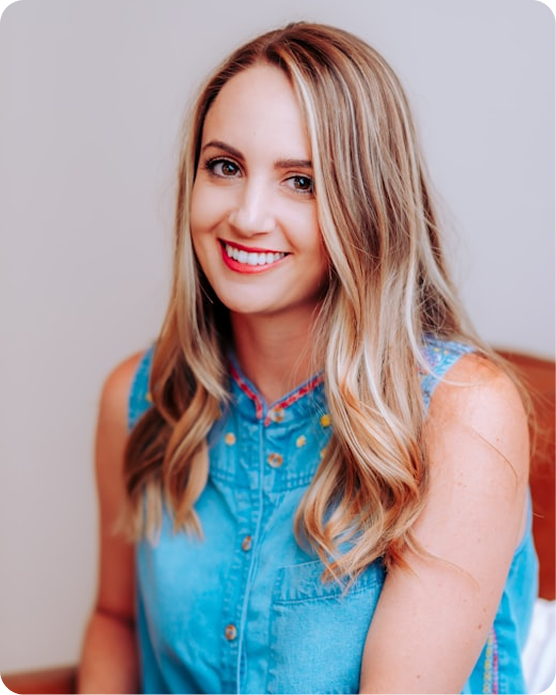 Smiling woman with long blonde hair wearing a blue sleeveless button-up shirt against a light background.