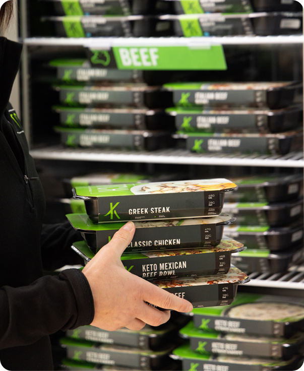 Person holding stacked keto meal trays labeled Greek Steak, Classic Chicken, Keto Mexican Beef Bowl, and Beef.