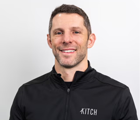 Smiling man with short dark hair wearing a black jacket with the word KITCH on it.