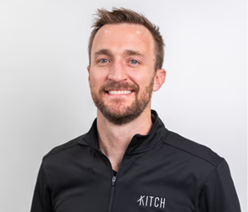 Smiling man with short brown hair and beard wearing a black Kitch jacket against a white background.