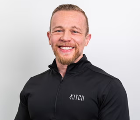 Smiling man in black zip-up with Kitch logo on chest against plain white background.