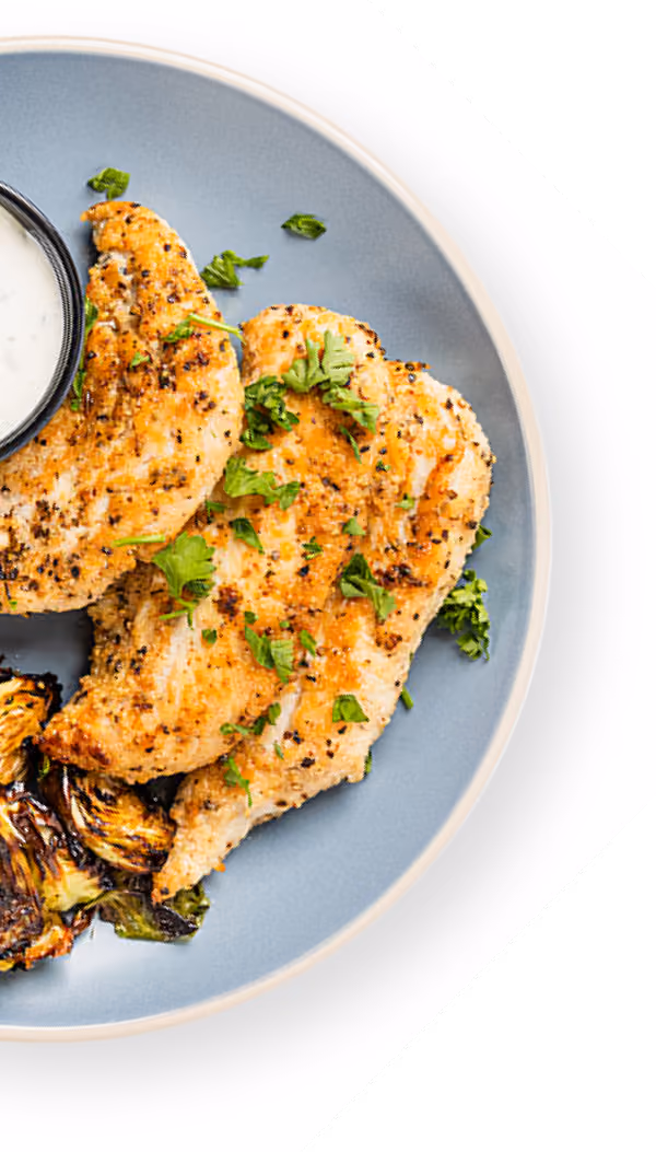 Grilled chicken breasts garnished with herbs on a blue plate with roasted vegetables and a side of white sauce.